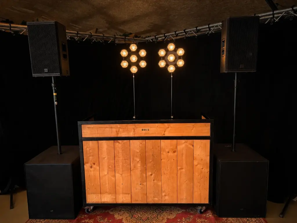 Houten DJ-booth met twee RCF KX-32A tops op subs en twee retrolampen, te huur bij dre-events